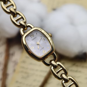 May include: A gold-tone Seiko Lucent wristwatch featuring a rectangular case with rounded corners and a mother-of-pearl dial. The watch has gold-tone hands and numerals at the 12, 3, 6, and 9 positions, attached to a thick, polished gold-tone link bracelet.
