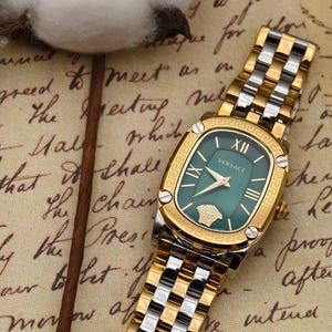 Puede incluir: Un reloj Versace dorado y plateado con esfera verde. El reloj tiene una esfera rectangular con n&uacute;meros romanos y un bisel dorado. La correa es una combinaci&oacute;n de eslabones dorados y plateados.
