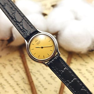 May include: A Seiko Credor wristwatch featuring a round silver-tone case and a gold-tone dial with thin black hour markers and hands. The watch includes a black leather strap with a textured crocodile-embossed pattern and a small crown on the side.