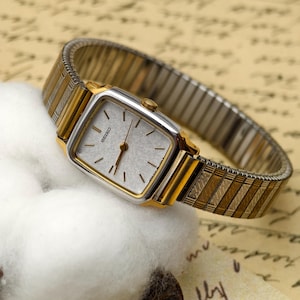 May include: A vintage Seiko watch with a square silver face and gold-toned bezel. The watch has gold and silver stretch band. The watch is resting on a white cotton ball. The watch face has the word "SEIKO" printed on it.