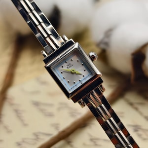 May include: A silver Vert Dense wristwatch with a square face and a light blue dial. The watch has a silver band and a small crown. The dial has a light blue background with silver dots and yellow hands. The text "JAPAN MOVT-921-9052848-A" is visible.