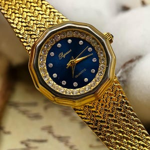 May include: Gold-toned Pegasus quartz watch with a blue face and a bezel encrusted with small, clear crystals. The watch has gold hands and a gold woven band. The watch face has the word "Pegasus" and "Quartz" printed on it.