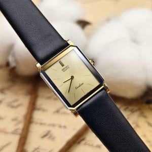 May include: A gold-toned Seiko Quartz Exceline watch with a rectangular face and black leather strap. The watch face is a light gold colour with black hands and the words "Seiko Quartz Exceline" printed on the face.
