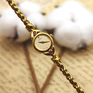 May include: A gold-tone Seiko wristwatch featuring a small round face with a textured cream dial, gold hands, and two small red stones set at the top and bottom of the case. The watch has a twisted rope-style metal link bracelet band.