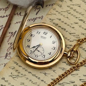 May include: A gold-tone Seiko Quartz pocket watch with a cream-colored dial, black Arabic numerals, and a sub-dial for seconds. The watch features a hinged cover and is attached to a matching gold-tone metal chain, displaying a classic round case.
