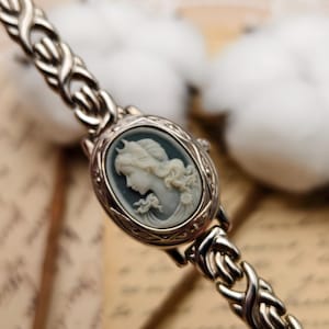 Puede incluir: Un reloj de pulsera plateado con una esfera ovalada que presenta un relieve de camafeo en azul claro y blanco de un retrato de perfil. La correa del reloj est&aacute; formada por eslabones met&aacute;licos plateados interconectados en un patr&oacute;n trenzado.
