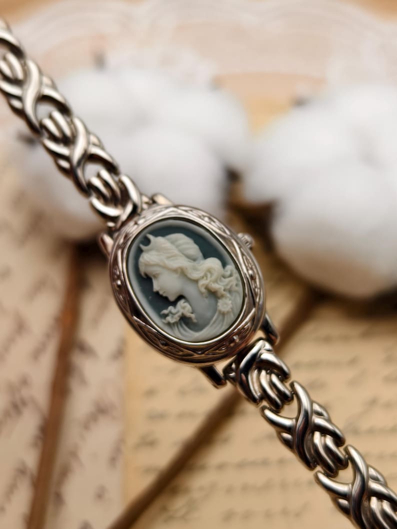 Puede incluir: Un reloj de pulsera plateado con una esfera ovalada que presenta un relieve de camafeo en azul claro y blanco de un retrato de perfil. La correa del reloj est&aacute; formada por eslabones met&aacute;licos plateados interconectados en un patr&oacute;n trenzado.