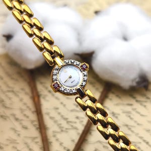 Puede incluir: Un reloj de pulsera Seiko en tono dorado con esfera redonda blanca, manecillas e índices dorados. El bisel está adornado con pequeñas piedras transparentes y dos piedras rojas cuadradas. El reloj tiene una pulsera de eslabones dorados.