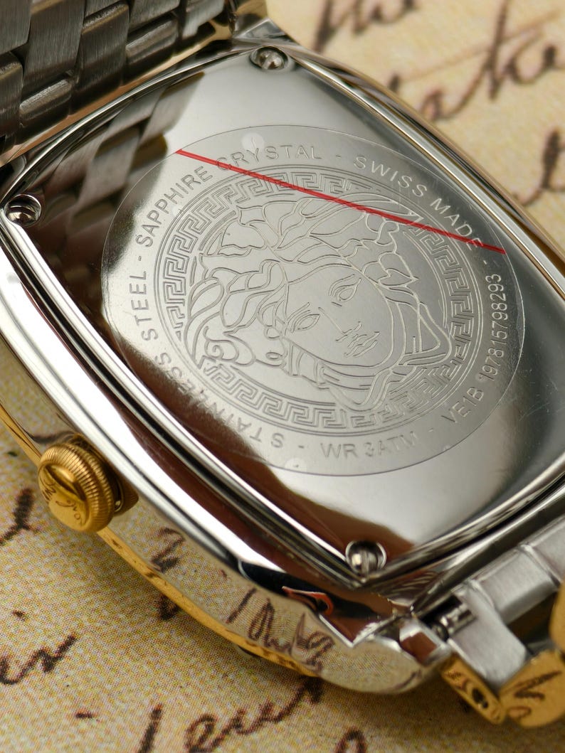 Puede incluir: Primer plano de un reloj de acero inoxidable con bisel y correa dorados. La parte trasera del reloj presenta un emblema de cabeza de Medusa y el texto "SAPPHIRE CRYSTAL SWISS MADE". Resistente al agua hasta 3 ATM.