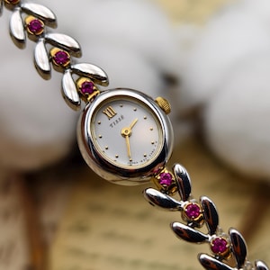 Puede incluir: Un reloj de pulsera plateado con esfera ovalada blanca que muestra el texto TISSÉ y un número romano XII. La correa consta de eslabones plateados en forma de hoja con pequeñas piedras redondas de color rosa en engastes dorados.