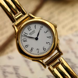 May include: A gold-plated Seiko Quartz wristwatch with a white face and black numerals. The watch has a gold strap and a classic design. The watch face displays the Seiko Quartz logo.