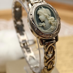Puede incluir: Un reloj de metal plateado con una esfera ovalada de camafeo. El camafeo muestra el perfil de una persona de color claro sobre un fondo gris oscuro. La correa del reloj consiste en un dise&ntilde;o intrincado de eslabones met&aacute;licos entrelazados.