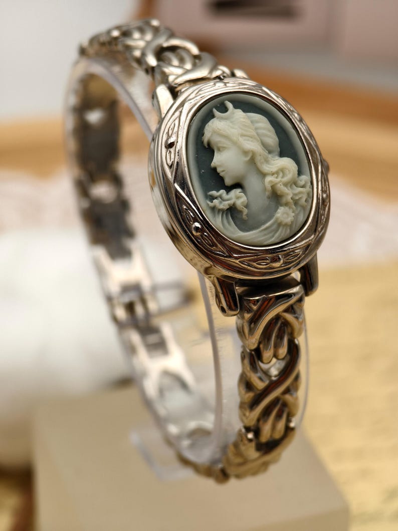 Puede incluir: Un reloj de metal plateado con una esfera ovalada de camafeo. El camafeo muestra el perfil de una persona de color claro sobre un fondo gris oscuro. La correa del reloj consiste en un dise&ntilde;o intrincado de eslabones met&aacute;licos entrelazados.