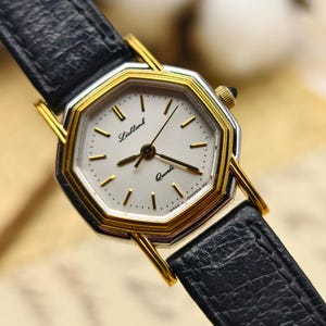 Könnte beinhalten: Eine Vintage-Armbanduhr mit einem schwarzen Lederarmband und einem goldfarbenen, achteckigen Gehäuse. Das Zifferblatt ist weiß mit goldenen Zeigern und Markierungen, und der Text "Lielbank Quartz" ist sichtbar. Das Armband ist etwa 1,3 cm breit.