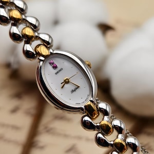 Può includere: Un orologio da polso Seiko Befina con quadrante ovale bianco, lancette color oro e due piccole pietre rosa in corrispondenza delle ore dodici. L'orologio presenta un cinturino a maglie bicolore argento e oro con finitura lucida e corona.