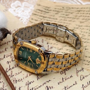 Puede incluir: Un reloj Versace dorado y plateado con una esfera ovalada verde. El reloj presenta n&uacute;meros romanos dorados, manecillas y un bisel dorado con un dise&ntilde;o de llave griega. La correa del reloj es una combinaci&oacute;n de eslabones dorados y plateados.
