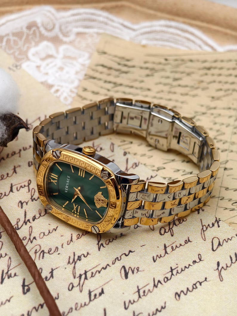 Puede incluir: Un reloj Versace dorado y plateado con una esfera ovalada verde. El reloj presenta n&uacute;meros romanos dorados, manecillas y un bisel dorado con un dise&ntilde;o de llave griega. La correa del reloj es una combinaci&oacute;n de eslabones dorados y plateados.