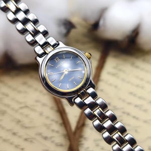 May include: A two-tone wristwatch featuring a round blue dial with gold-tone hands and hour markers. The watch face displays the text LUCENT and SEIKO. It has a silver and gold-tone metal link bracelet with a polished finish and a small gold crown.