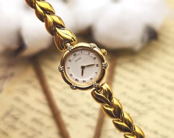 May include: A gold-tone wristwatch with a braided link bracelet and a round white face featuring the text TISSÉ. The bezel is accented with small clear stones at the hour markers, and the dial displays black Roman numeral indices.