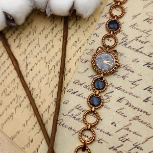 Może przedstawiać: Zegarek w stylu vintage w odcieniach złota z ciemnoszarą tarczą i złotymi wskaz&oacute;wkami. Pasek zegarka składa się z szeregu połączonych k&oacute;ł i ciemnoniebieskich owalnych akcent&oacute;w. Zegarek jest prezentowany na tle postarzanego papieru i łodyg bawełny.