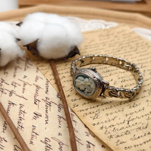 Puede incluir: Un reloj de pulsera plateado con una esfera ovalada que presenta un relieve de camafeo en azul claro y blanco con un retrato de perfil. El reloj tiene una intrincada pulsera de eslabones met&aacute;licos y una peque&ntilde;a corona en el lateral de la caja.
