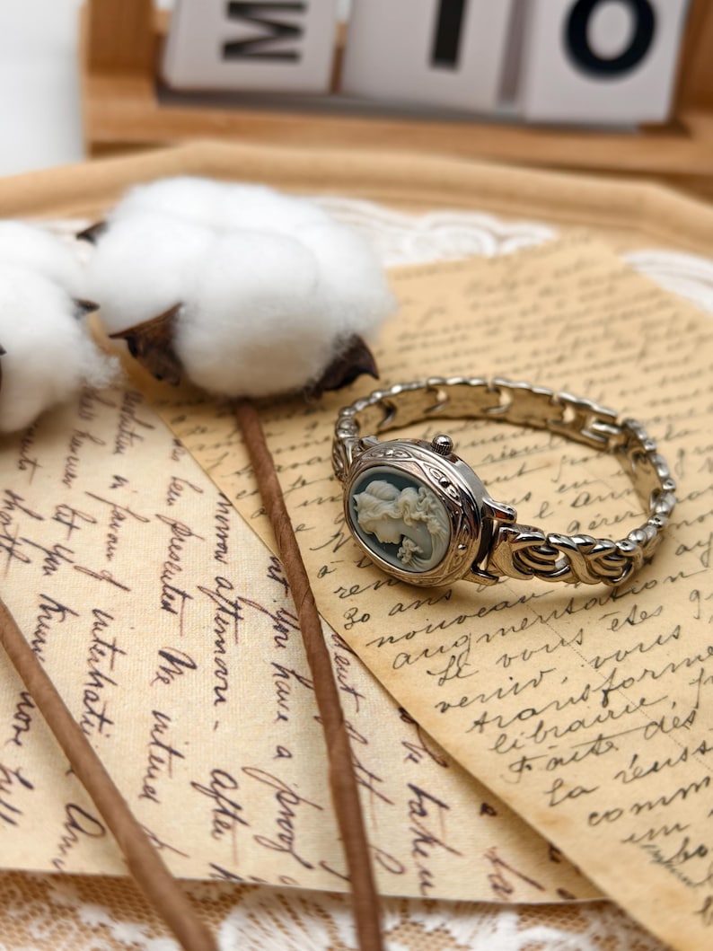 Puede incluir: Un reloj de pulsera plateado con una esfera ovalada que presenta un relieve de camafeo en azul claro y blanco con un retrato de perfil. El reloj tiene una intrincada pulsera de eslabones met&aacute;licos y una peque&ntilde;a corona en el lateral de la caja.