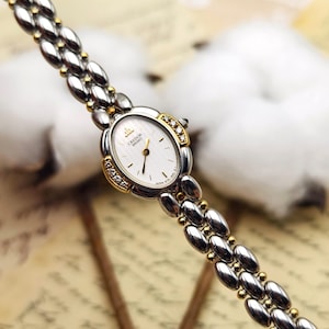 May include: A Credor Seiko wristwatch featuring an oval white dial with gold hands and indices. The bezel is accented with small clear stones on the sides. The bracelet is constructed from polished silver-tone oval links connected by small gold-tone beads.
