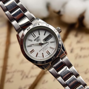 Puede incluir: Un reloj de pulsera Seiko plateado con esfera blanca, manecillas y marcadores negros. El reloj tiene una pantalla de fecha y una correa de acero inoxidable. La esfera del reloj dice "WATER SOM RESIST AUTOMATIC 21 JEWELS MADE IN JAPAN".