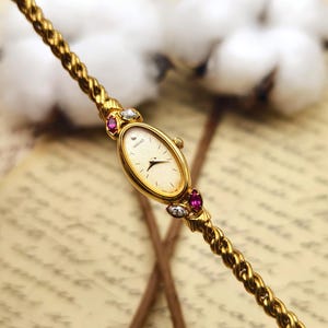 May include: A gold-tone Seiko wristwatch with an oval face and a textured cream dial. The watch features a twisted rope-style chain band accented with small, marquise-cut red stones and clear, round-cut stones flanking the watch case on both sides.