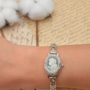 Puede incluir: Un reloj de pulsera plateado con una esfera de camafeo ovalada, fondo azul claro y un relieve de perfil blanco. El reloj cuenta con una correa de eslabones met&aacute;licos texturizados y una peque&ntilde;a corona en el lado derecho de la caja del reloj.