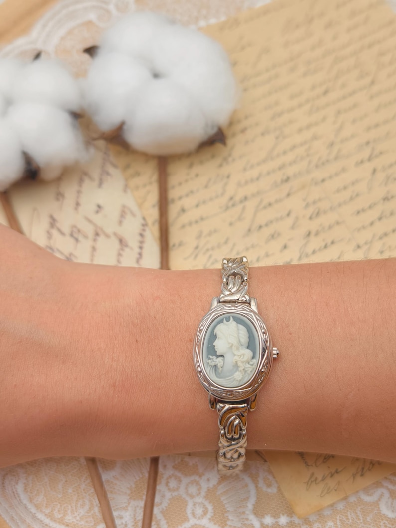 Puede incluir: Un reloj de pulsera plateado con una esfera de camafeo ovalada, fondo azul claro y un relieve de perfil blanco. El reloj cuenta con una correa de eslabones met&aacute;licos texturizados y una peque&ntilde;a corona en el lado derecho de la caja del reloj.