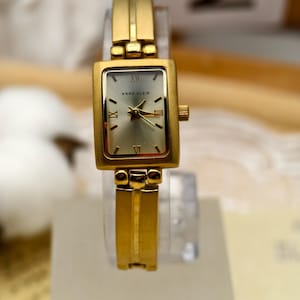 May include: A gold-coloured Anne Klein wristwatch with a rectangular face and Roman numeral hour markers. The watch has a gold band and a silver-coloured watch face. The watch is displayed on a clear stand.