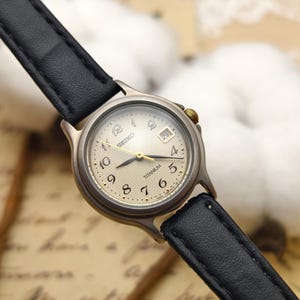 Könnte beinhalten: Eine silberfarbene Seiko-Uhr mit schwarzem Lederarmband. Das cremefarbene Zifferblatt hat goldene Zeiger und Ziffern. Eine Datumsanzeige befindet sich auf 3 Uhr. Das Wort "Titanium" ist auf dem Zifferblatt sichtbar.