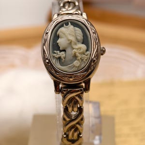 Puede incluir: Un reloj de pulsera dorado con una esfera ovalada que presenta un retrato de camafeo en gris y blanco de una figura de perfil. El reloj tiene una correa de eslabones met&aacute;licos entrelazados y una peque&ntilde;a corona en el lateral derecho.