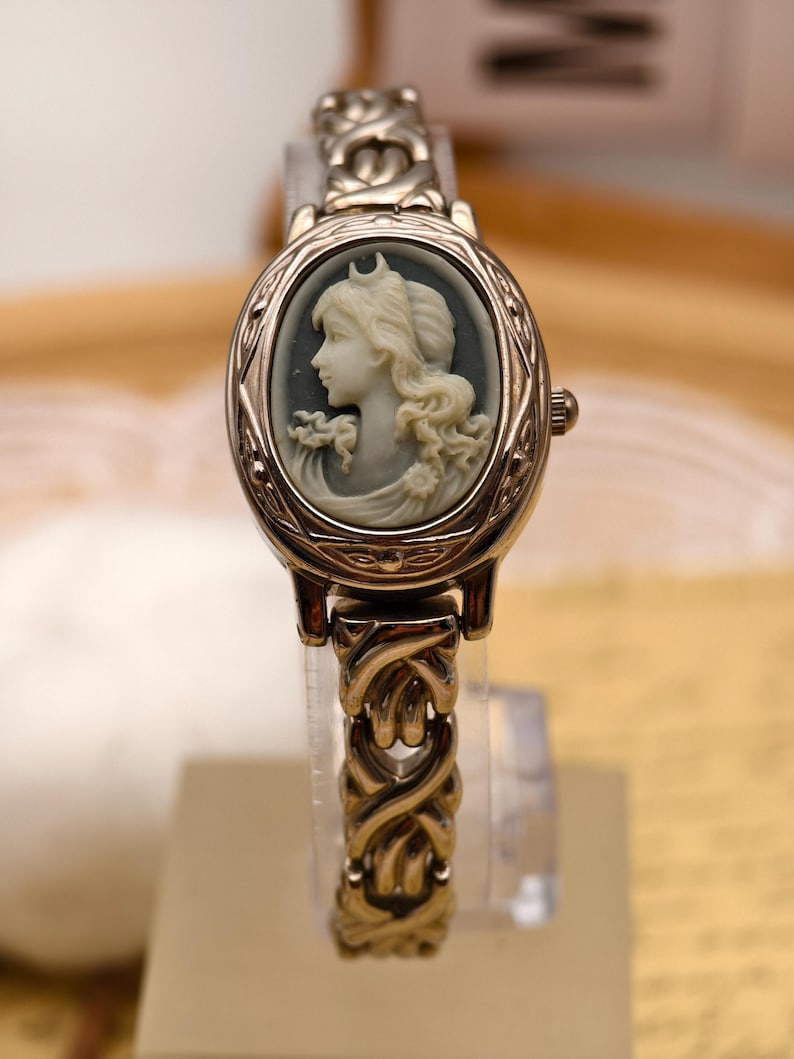 Puede incluir: Un reloj de pulsera dorado con una esfera ovalada que presenta un retrato de camafeo en gris y blanco de una figura de perfil. El reloj tiene una correa de eslabones met&aacute;licos entrelazados y una peque&ntilde;a corona en el lateral derecho.