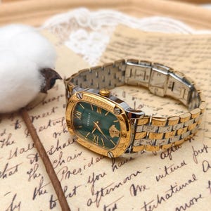 Puede incluir: Un reloj Versace dorado y plateado con esfera verde. El reloj tiene n&uacute;meros romanos y un bisel dorado. El reloj est&aacute; sobre un fondo de papel vintage.