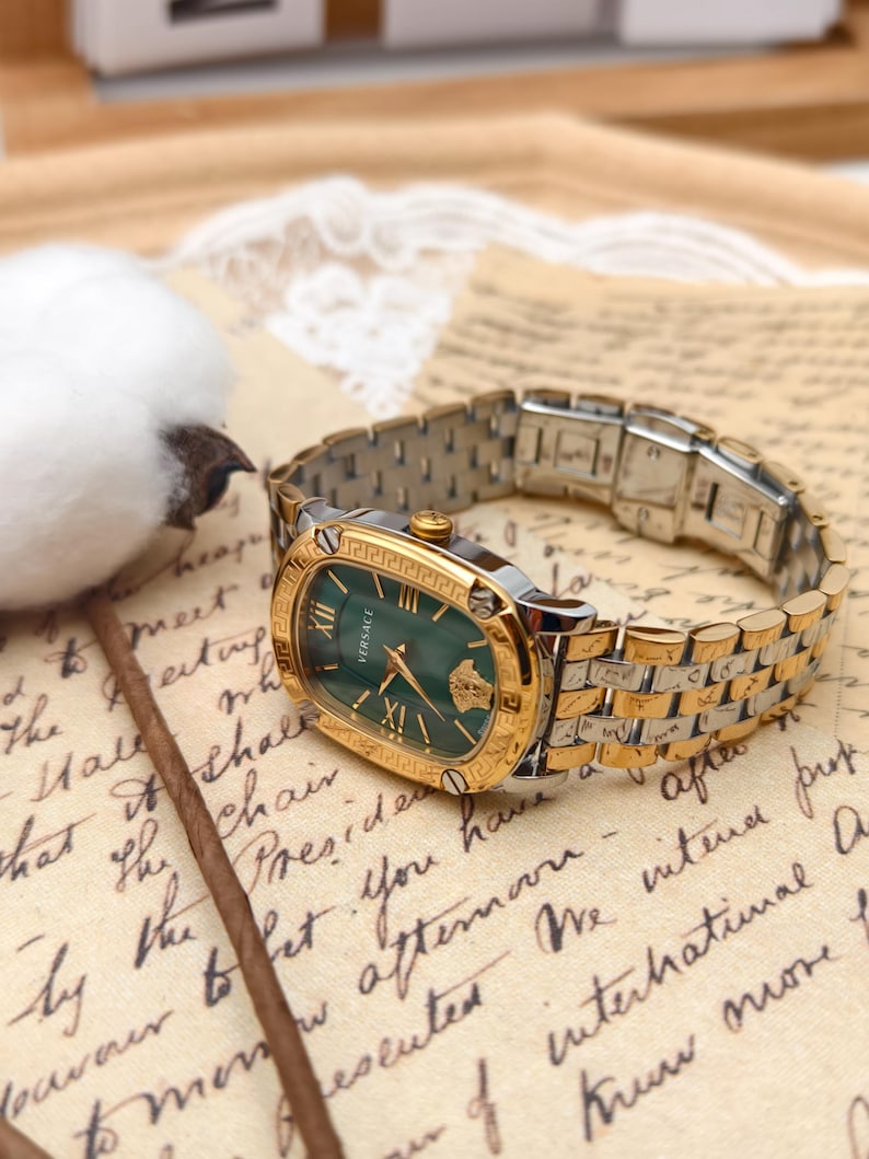 Puede incluir: Un reloj Versace dorado y plateado con esfera verde. El reloj tiene n&uacute;meros romanos y un bisel dorado. El reloj est&aacute; sobre un fondo de papel vintage.