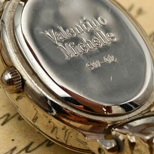Puede incluir: Un reloj de pulsera de metal plateado con una tapa trasera ovalada. La parte posterior est&aacute; grabada con el texto Valentino Michelle y el n&uacute;mero de modelo SW-02. El reloj cuenta con una peque&ntilde;a corona lateral y asas met&aacute;licas.