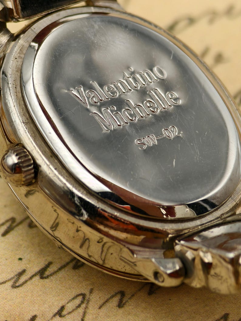 Puede incluir: Un reloj de pulsera de metal plateado con una tapa trasera ovalada. La parte posterior est&aacute; grabada con el texto Valentino Michelle y el n&uacute;mero de modelo SW-02. El reloj cuenta con una peque&ntilde;a corona lateral y asas met&aacute;licas.