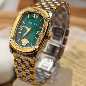 Puede incluir: Reloj Versace dorado y plateado con esfera verde. El reloj presenta n&uacute;meros romanos, agujas doradas y un bisel dorado con dise&ntilde;o de llave griega. La correa es una combinaci&oacute;n de eslabones dorados y plateados.