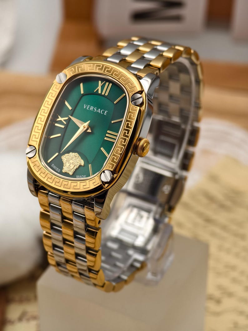 Puede incluir: Reloj Versace dorado y plateado con esfera verde. El reloj presenta n&uacute;meros romanos, agujas doradas y un bisel dorado con dise&ntilde;o de llave griega. La correa es una combinaci&oacute;n de eslabones dorados y plateados.