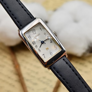 May include: A rectangular Seiko wristwatch featuring a silver-tone case with gold-tone accents, a white dial with black Arabic numerals, and thin blue hands. The watch is attached to a slim, black leather strap with visible black stitching.