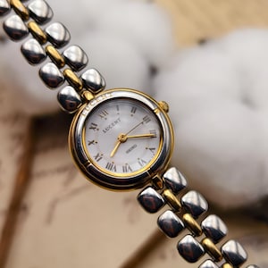 Könnte beinhalten: Gold-silberne Lucent Seiko-Uhr mit weißem Zifferblatt und römischen Ziffern. Die Uhr hat eine goldene Lünette und ein silber-goldenes Gliederarmband. Ein klassisches, elegantes Design.
