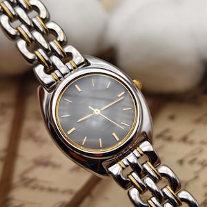 May include: A silver and gold-toned Seiko Lucent wristwatch with a dark grey face. The watch features gold hands and hour markers, and a linked bracelet. The watch is displayed against a soft, blurred background.