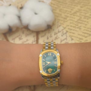 Puede incluir: Un reloj Versace dorado y plateado con esfera verde se muestra en una mu&ntilde;eca. El reloj presenta n&uacute;meros romanos y el logotipo de Versace. La correa del reloj es una combinaci&oacute;n de eslabones dorados y plateados.