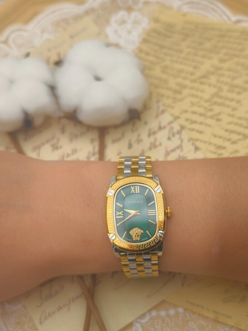 Puede incluir: Un reloj Versace dorado y plateado con esfera verde se muestra en una mu&ntilde;eca. El reloj presenta n&uacute;meros romanos y el logotipo de Versace. La correa del reloj es una combinaci&oacute;n de eslabones dorados y plateados.