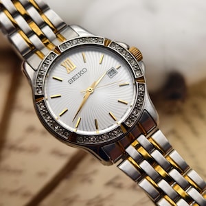 Puede incluir: Reloj Seiko plateado y dorado con esfera blanca y visualización de fecha. El reloj tiene un bisel con diamantes y agujas e indicadores dorados. La correa es una combinación de eslabones plateados y dorados.