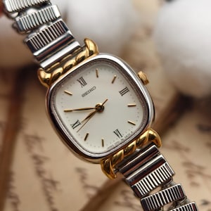 May include: A vintage Seiko watch with a square, white face and gold accents. The watch features Roman numeral hour markers and gold hands. The watch band is silver and gold, with a flexible, expandable design. The watch is a classic timepiece.