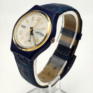 Puede incluir: Un reloj Swatch Swiss con caja azul marino y bisel dorado. La esfera es blanquecina con números romanos y agujas doradas. La correa es de cuero azul marino. La fecha indica "DOM 14".