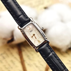 May include: A rectangular Credor Seiko wristwatch featuring a silver-toned case, a white textured dial with gold-colored hands, and Roman numeral markers at the twelve and six o'clock positions. The watch includes a black leather strap with a crocodile-embossed pattern.